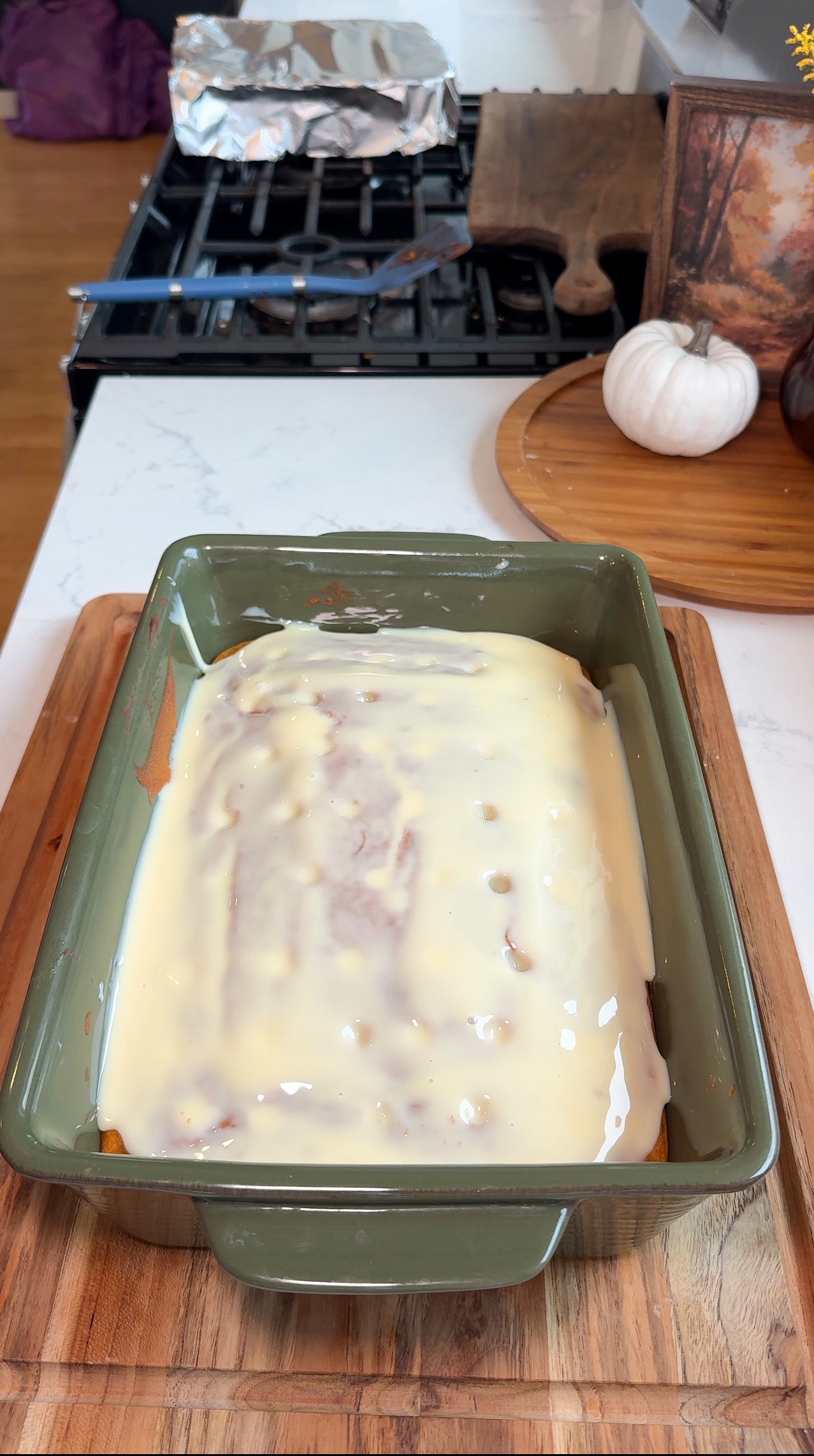 A green baking dish filled with food covered in a creamy white sauce sits on a wooden cutting board in a kitchen, with a white pumpkin and other kitchen items in the background.