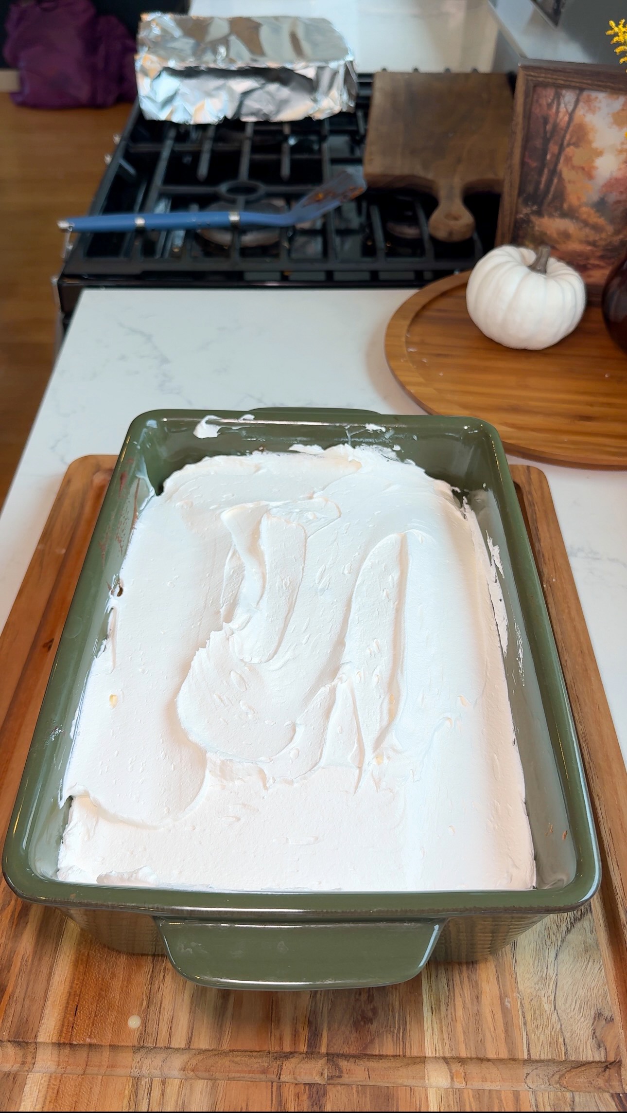 A green baking dish filled with a dessert covered in smooth white whipped topping sits on a wooden cutting board in a kitchen.