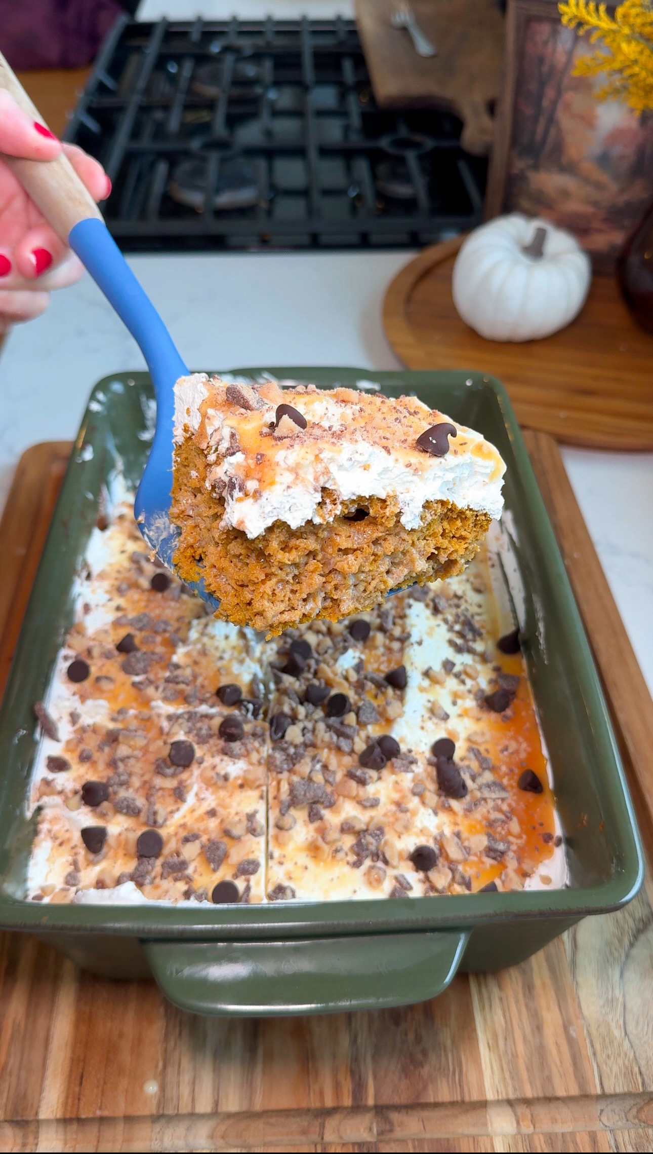 A hand holds a spatula lifting a piece of pumpkin cake topped with whipped cream, caramel, chocolate chips, and toffee bits from a green baking dish on a wooden board.