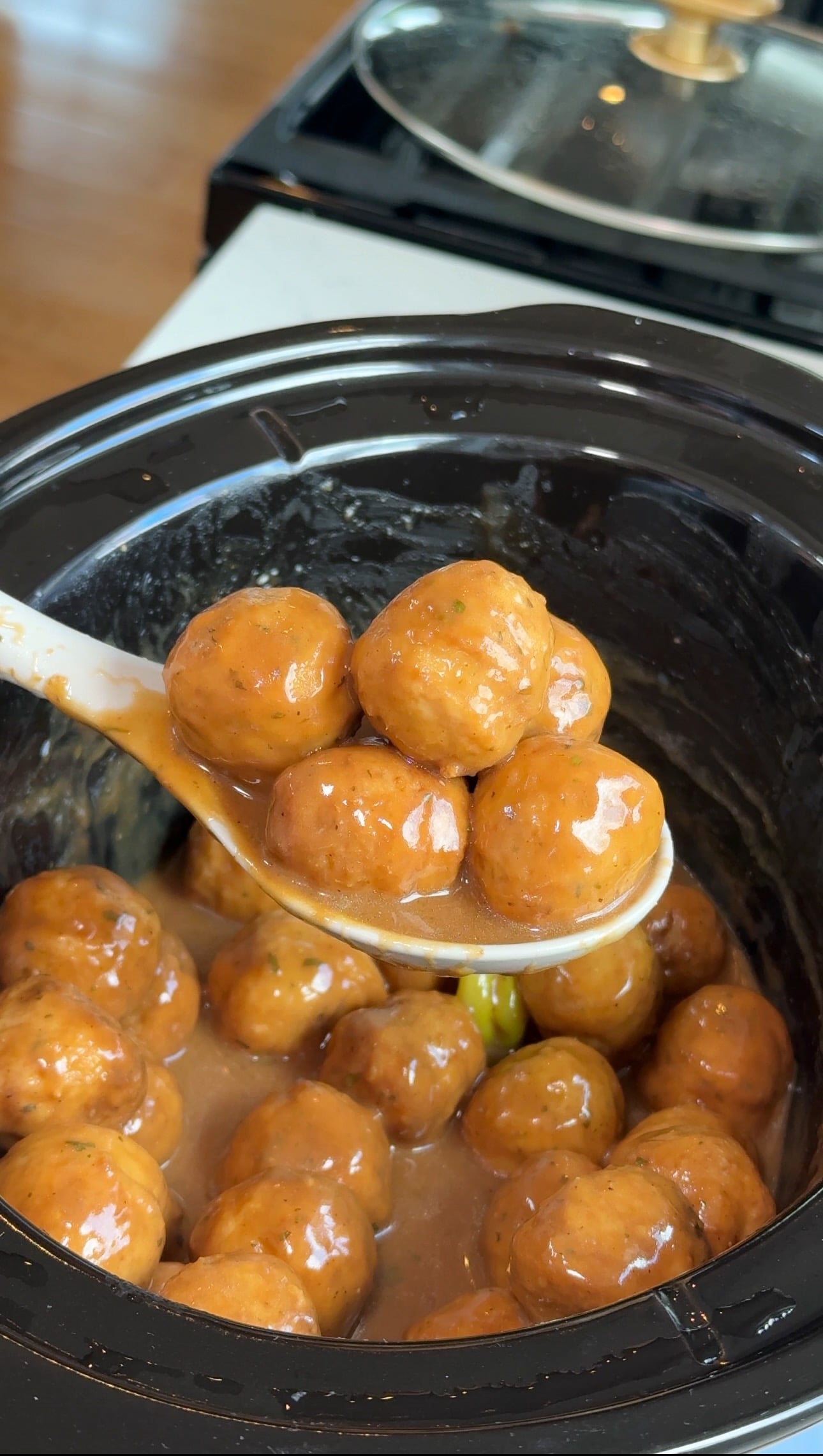 A white spoon lifts several saucy meatballs from a black slow cooker filled with more meatballs and sauce.