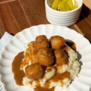 A white plate with mashed potatoes topped with gravy and meatballs, garnished with herbs. In the background, a small bowl of yellow peppers sits on a wooden tray. Sunlight brightens the scene.