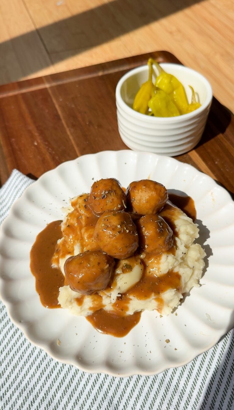 A white plate with mashed potatoes topped with gravy and meatballs, garnished with herbs. In the background, a small bowl of yellow peppers sits on a wooden tray. Sunlight brightens the scene.
