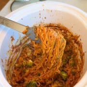 A fork lifts a serving of saucy noodles with vegetables from a white slow cooker, showing a mix of noodles, sauce, and broccoli inside.
