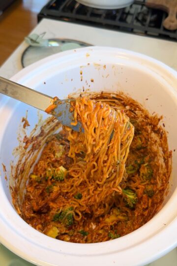A fork lifts a serving of saucy noodles with vegetables from a white slow cooker, showing a mix of noodles, sauce, and broccoli inside.
