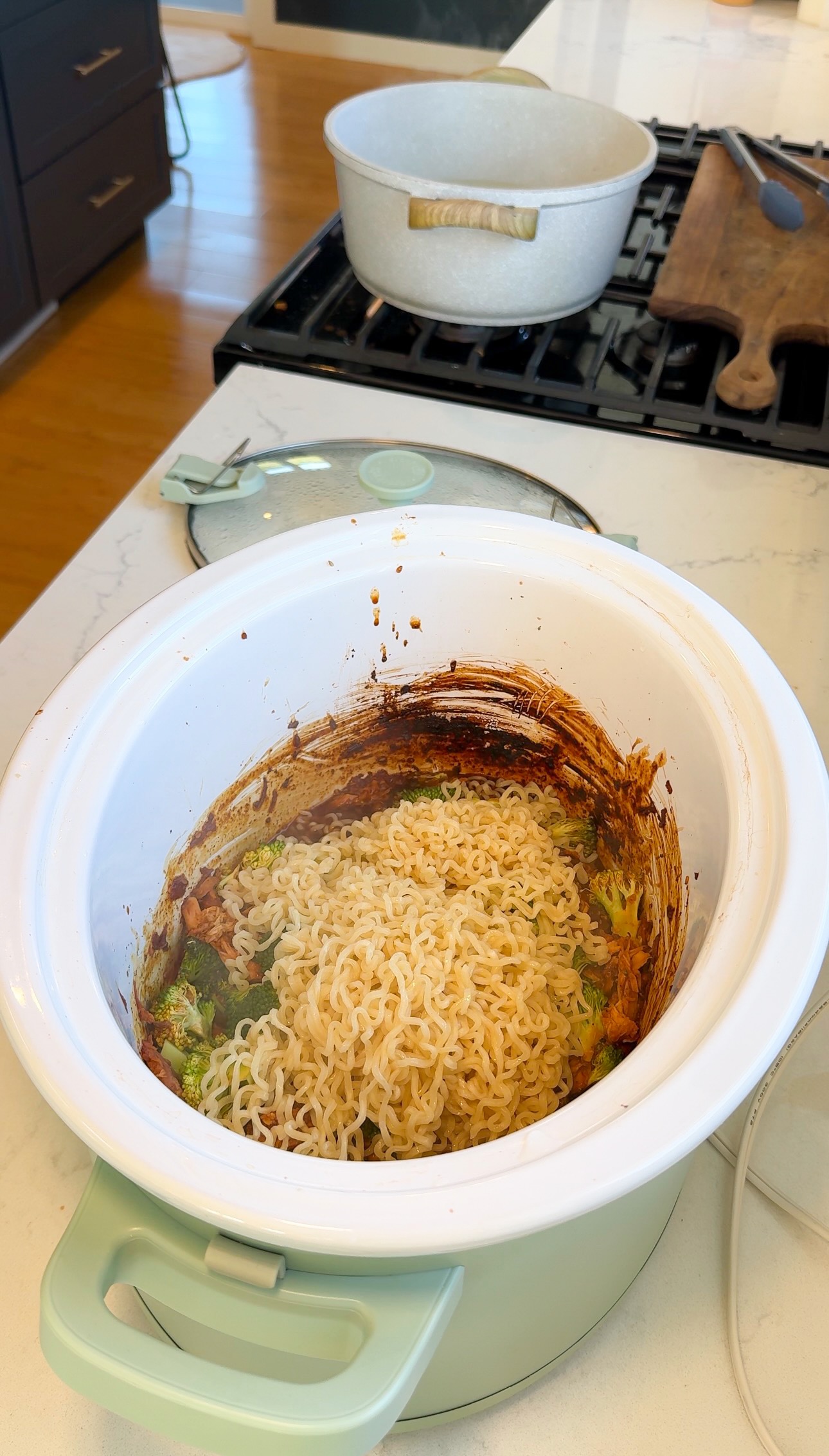 A slow cooker with cooked instant noodles, sauce residue on the sides, and vegetables sits on a kitchen counter; a pot and wooden cutting board are in the background on the stove.