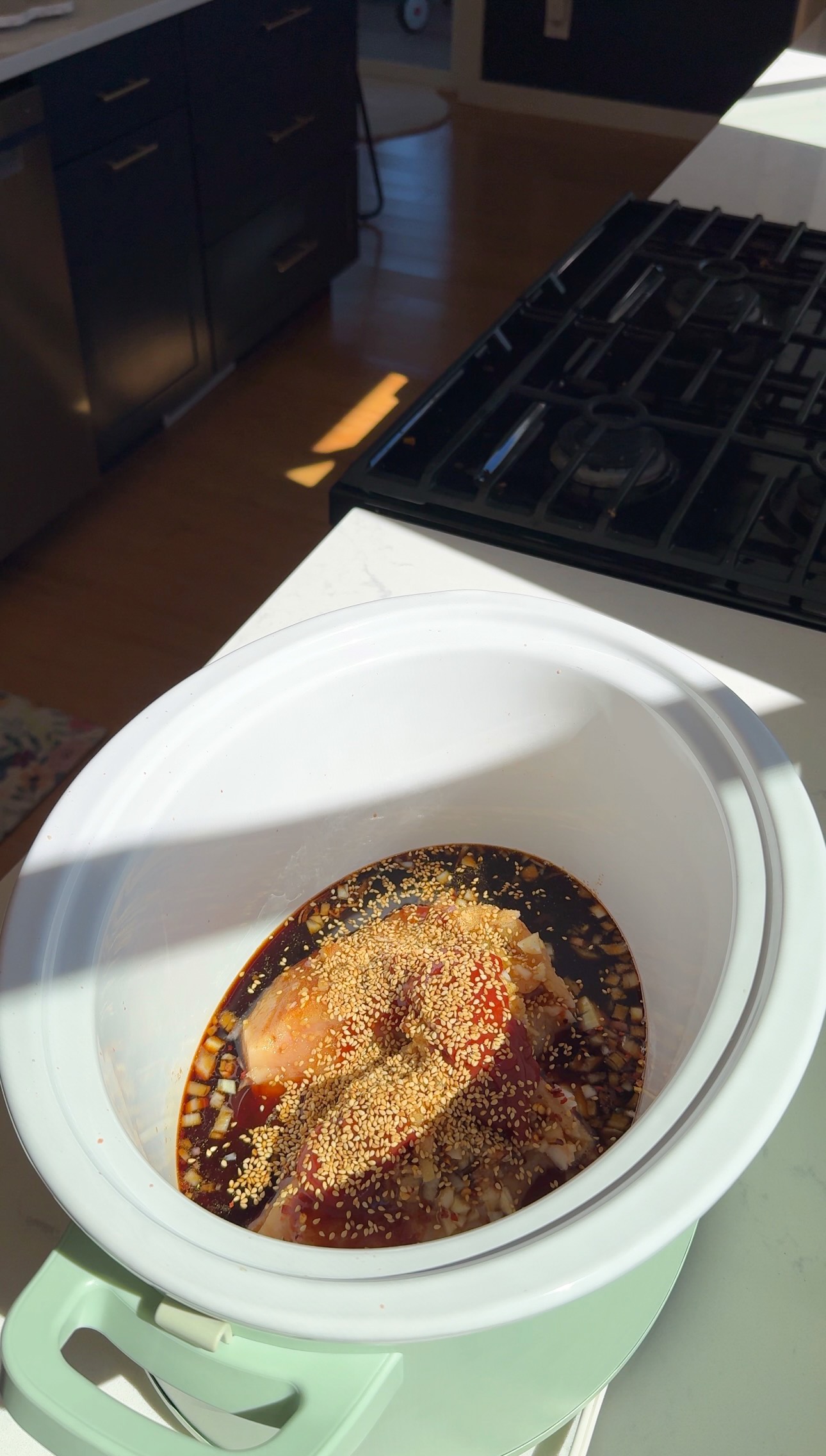 Raw chicken covered in sauce, spices, and sesame seeds sits in a white slow cooker on a kitchen counter, with sunlight streaming in from the left.