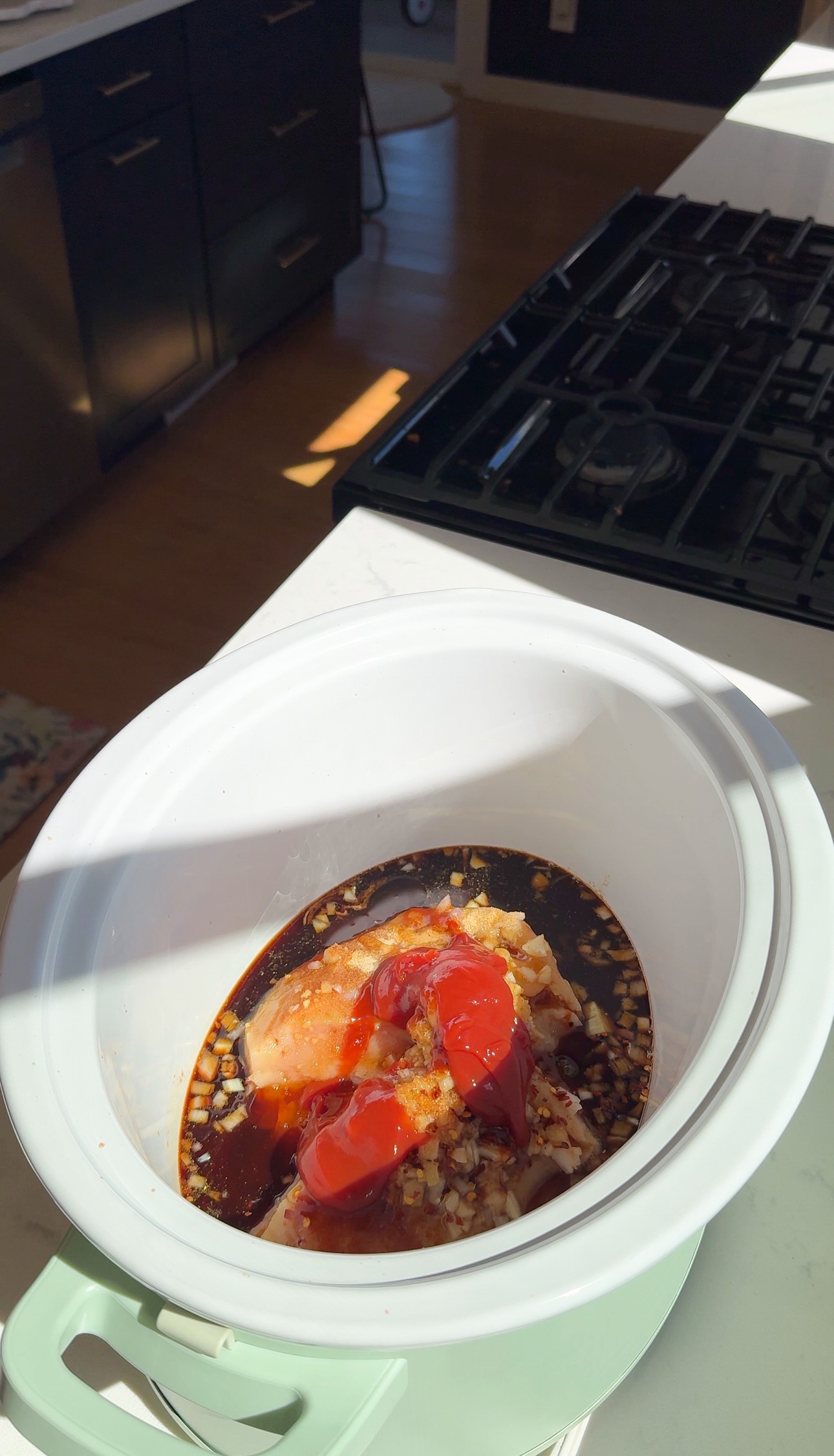 Raw chicken breasts in a slow cooker topped with chopped garlic, soy sauce, and ketchup, sitting on a white kitchen counter with sunlight streaming in.