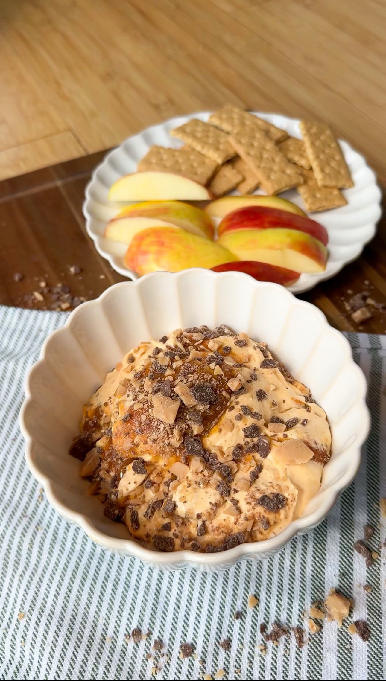 A bowl of creamy dip topped with crushed toffee sits on a striped cloth, with a plate of sliced apples and graham crackers in the background on a wooden surface.