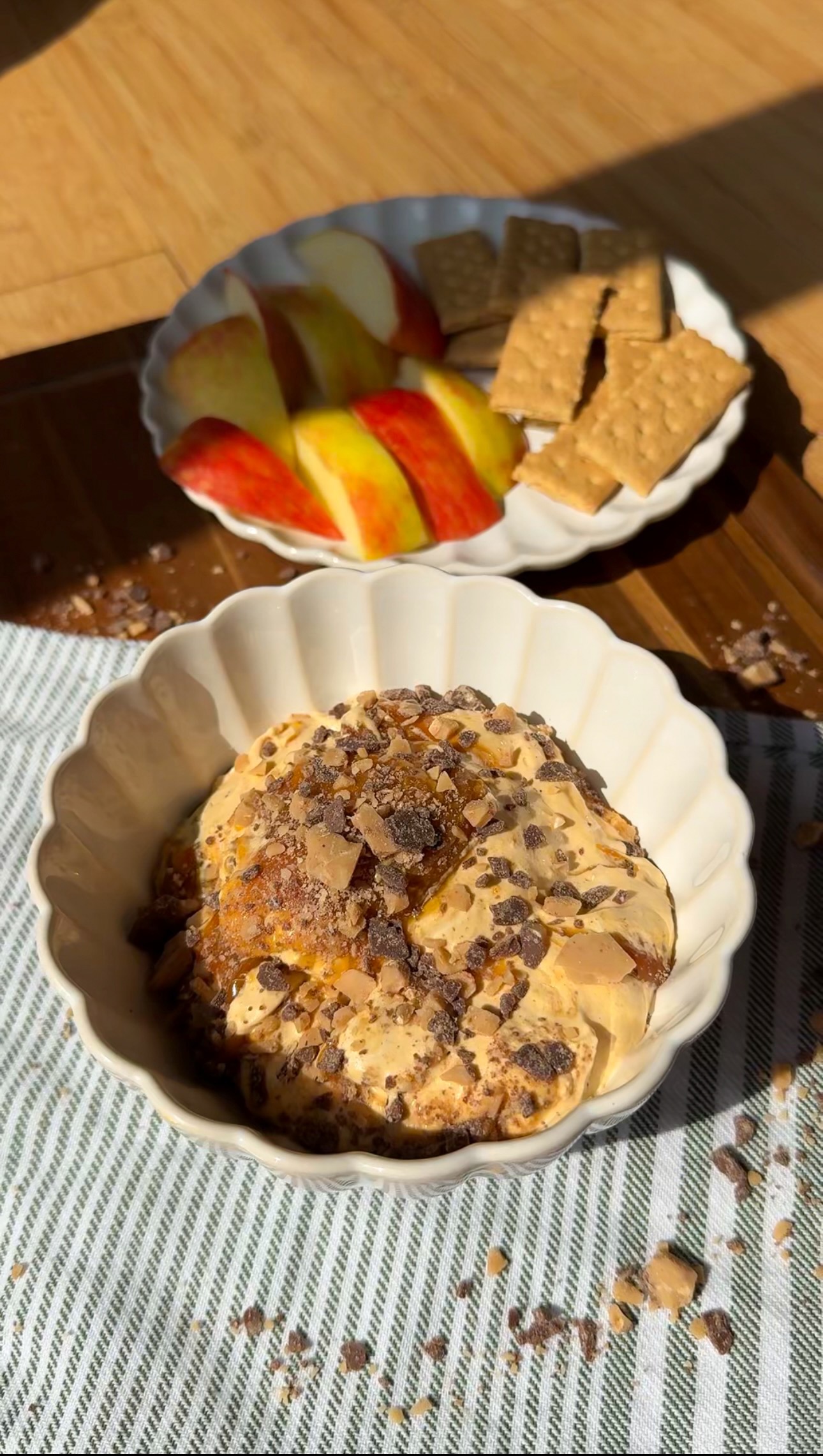 Bowl of dessert topped with chocolate shavings in the foreground. Behind it, a plate with apple slices and graham crackers sits on a wooden surface, sunlight streaming in.