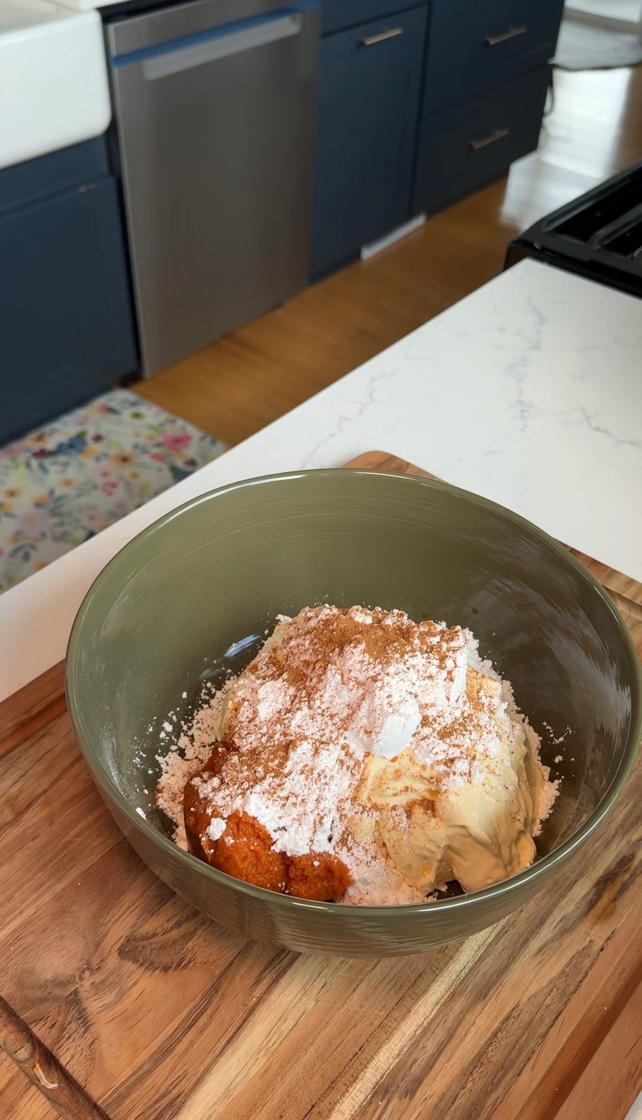 A green bowl on a kitchen counter holds cream cheese, pumpkin puree, powdered sugar, and spices, ready to be mixed. The background shows cabinets, a stove, and a floral rug on the floor.