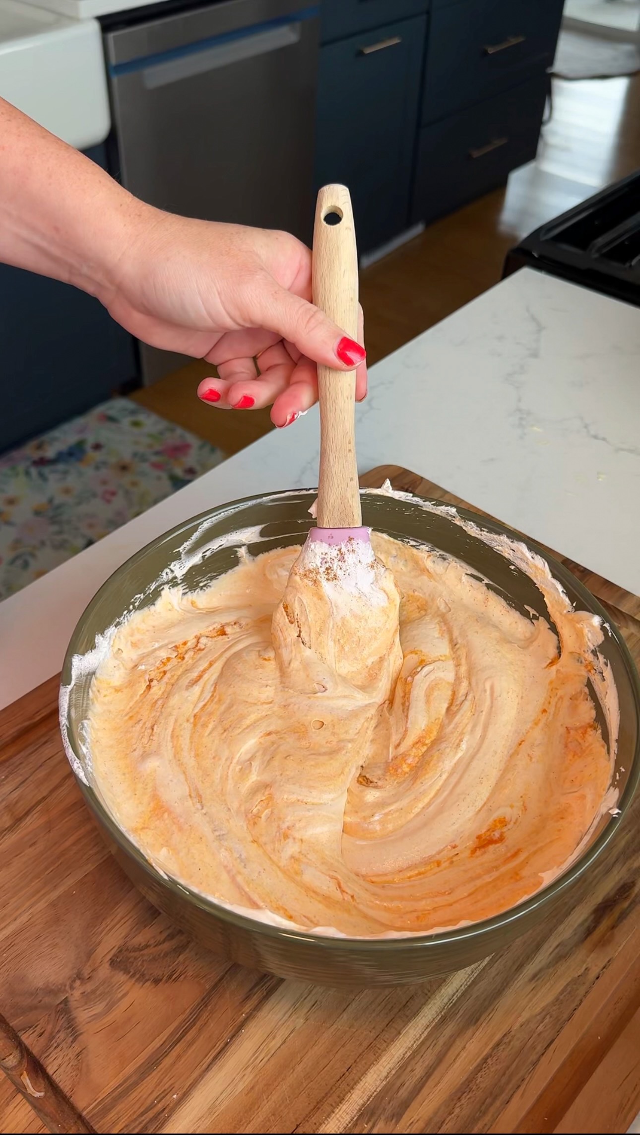 A hand with red nail polish holds a spatula, stirring an orange and white creamy mixture in a glass bowl on a kitchen counter.