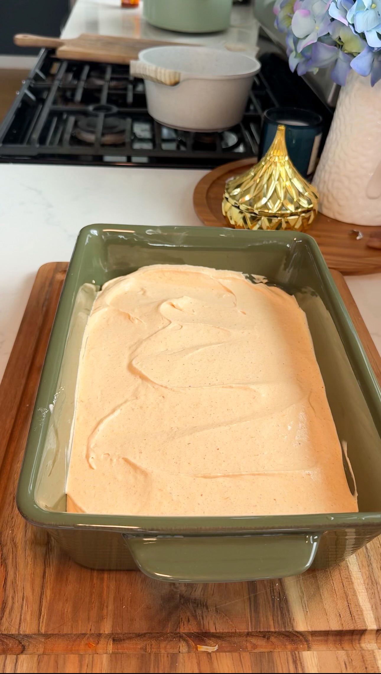 A green rectangular baking dish filled with a smooth, light orange batter sits on a wooden board. In the background are a stove, a white canister, a gold decorative object, and blue flowers.