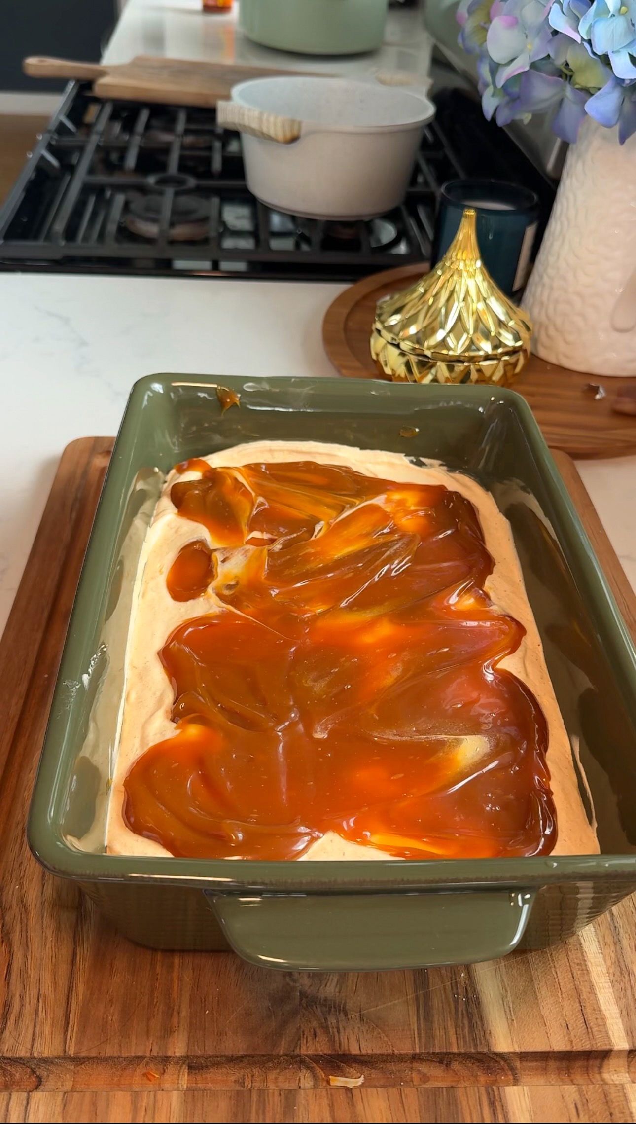A rectangular green baking dish filled with a creamy dessert, topped with swirled caramel sauce, sits on a wooden board in a kitchen setting. Decorative items and a stove are visible in the background.