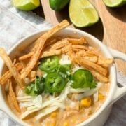 A bowl of creamy soup topped with shredded cheese, tortilla strips, jalapeño slices, cilantro, and corn, garnished with lime wedges and jalapeños in the background.