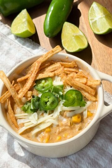 A bowl of creamy soup topped with shredded cheese, tortilla strips, jalapeño slices, cilantro, and corn, garnished with lime wedges and jalapeños in the background.