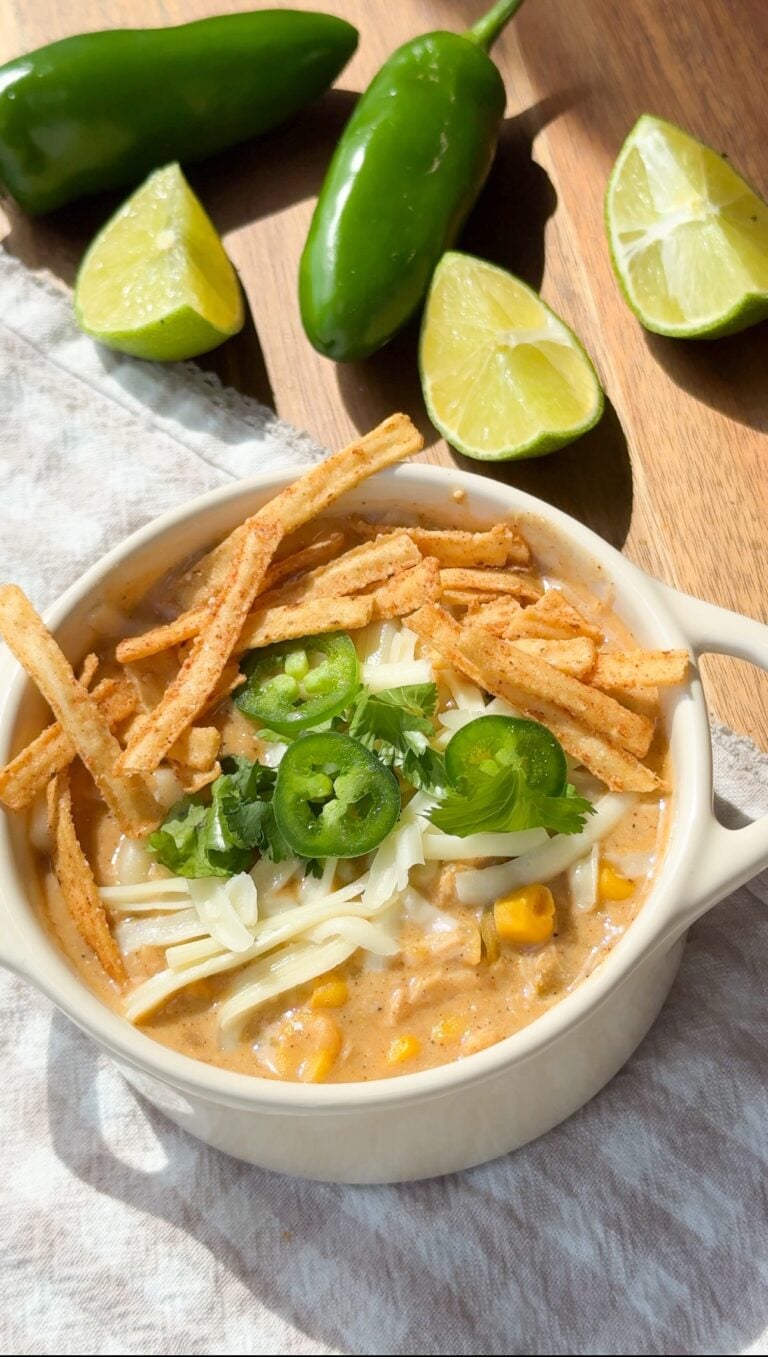 A bowl of creamy soup topped with shredded cheese, tortilla strips, jalapeño slices, cilantro, and corn, garnished with lime wedges and jalapeños in the background.