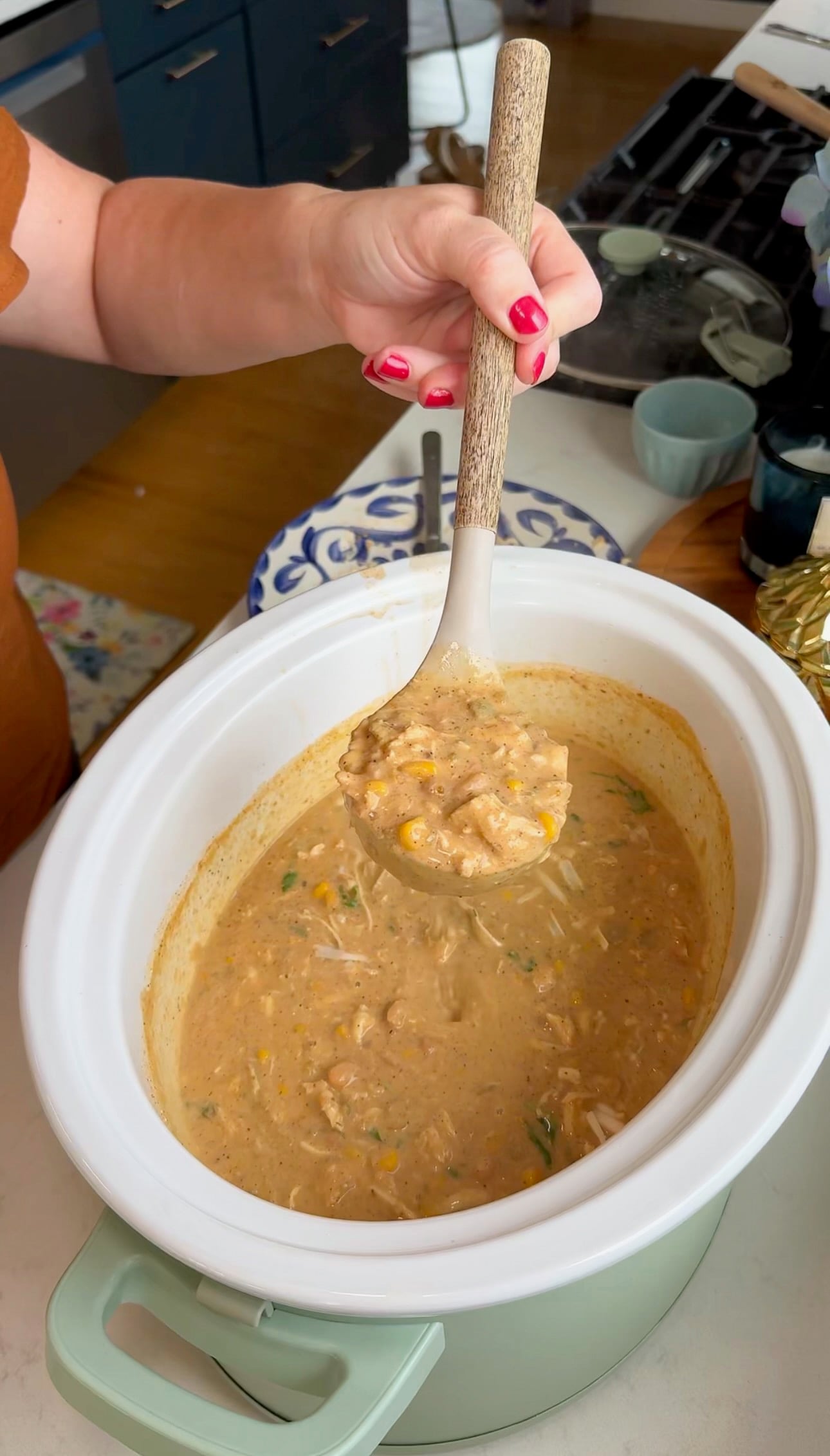 A person holding a ladle of thick, creamy soup with vegetables and beans over a white slow cooker in a kitchen.