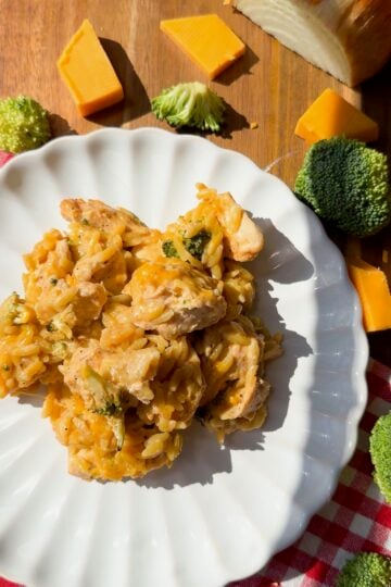 A plate of chicken broccoli orzo casserole with melted cheddar, surrounded by broccoli florets, cheddar cheese, and an onion.