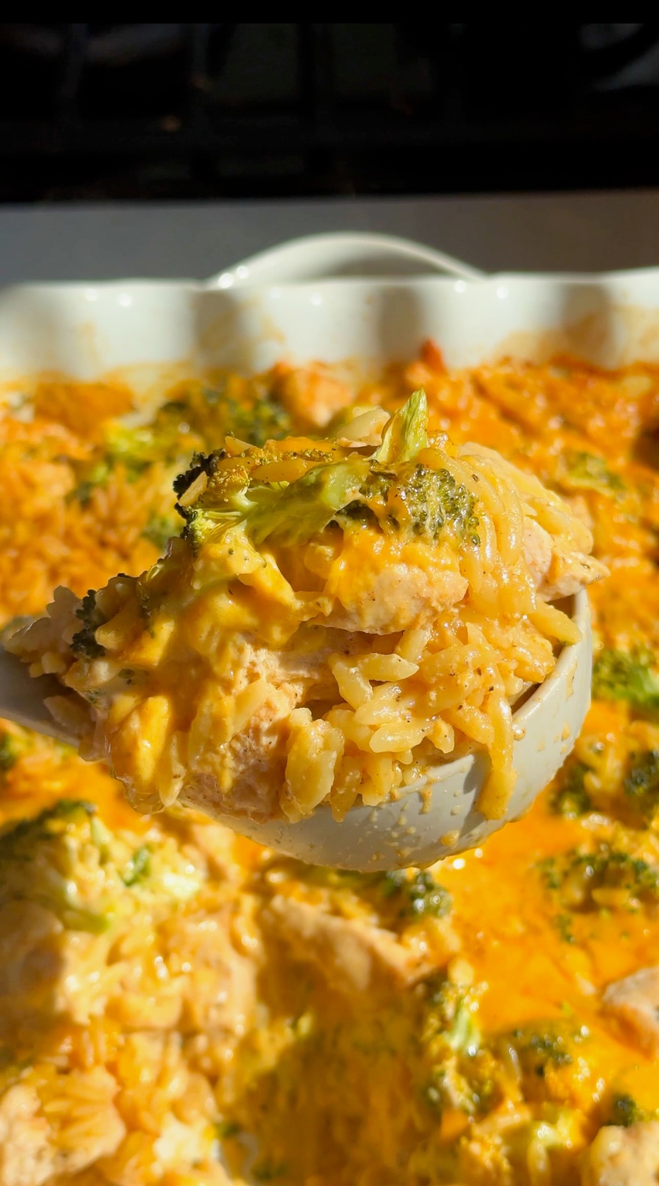 A serving spoon lifting a scoop of cheesy chicken broccoli orzo from the casserole dish.