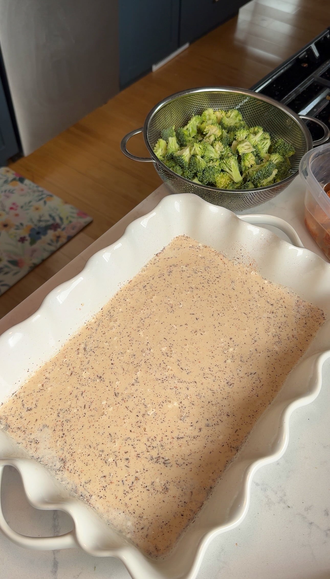 Creamy seasoned chicken stock and heavy cream whisked together in a white 9x13 baking dish, with diced broccoli in a colander nearby.
