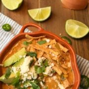 A bowl of creamy chicken tortilla soup topped with avocado, queso fresco, cilantro, and sliced jalapeño, with lime wedges nearby.