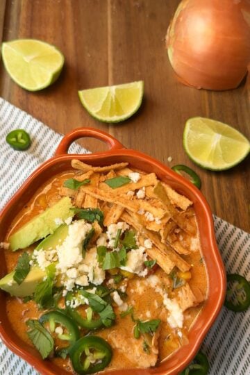 A bowl of creamy chicken tortilla soup topped with avocado, queso fresco, cilantro, and sliced jalapeño, with lime wedges nearby.