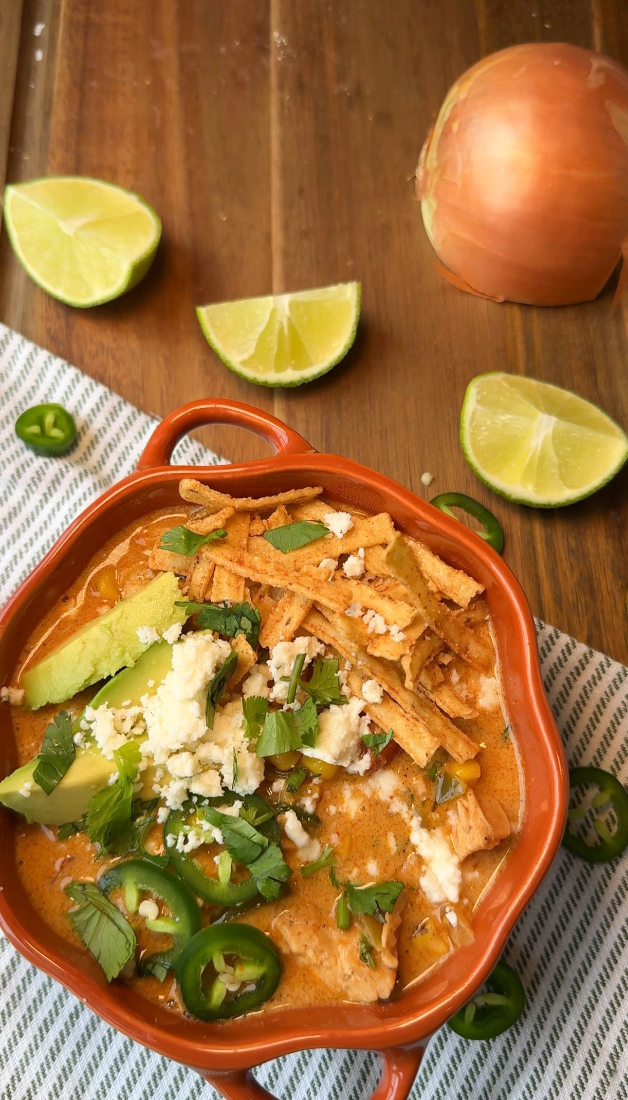 A bowl of creamy chicken tortilla soup topped with avocado, queso fresco, cilantro, and sliced jalapeño, with lime wedges nearby.