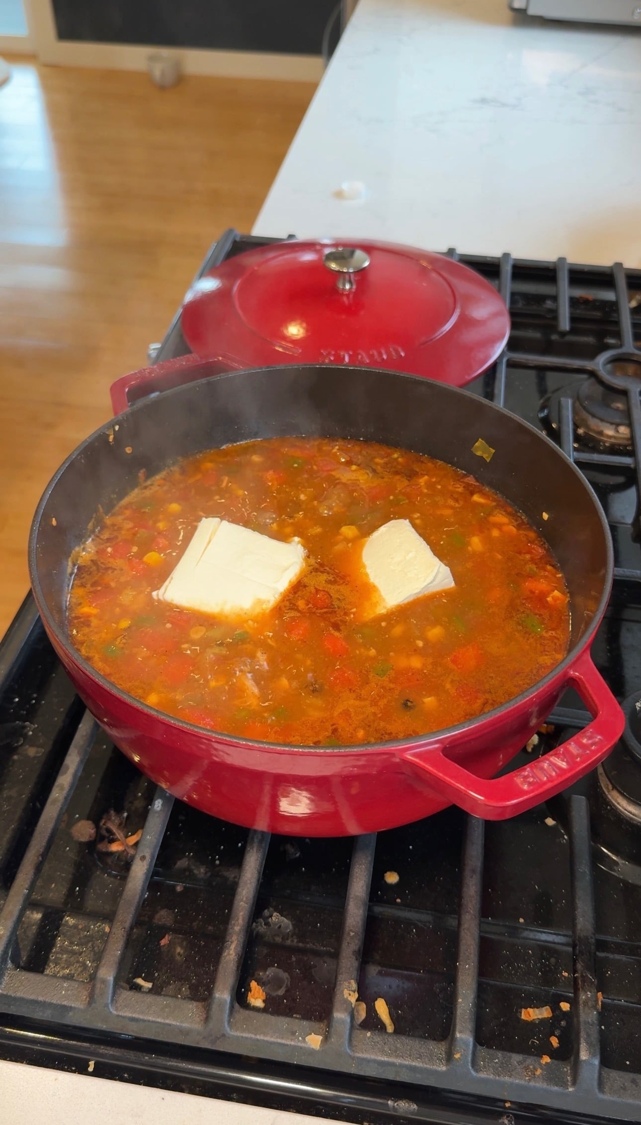 Two blocks of softened cream cheese melting into chicken tortilla soup in a red pot.