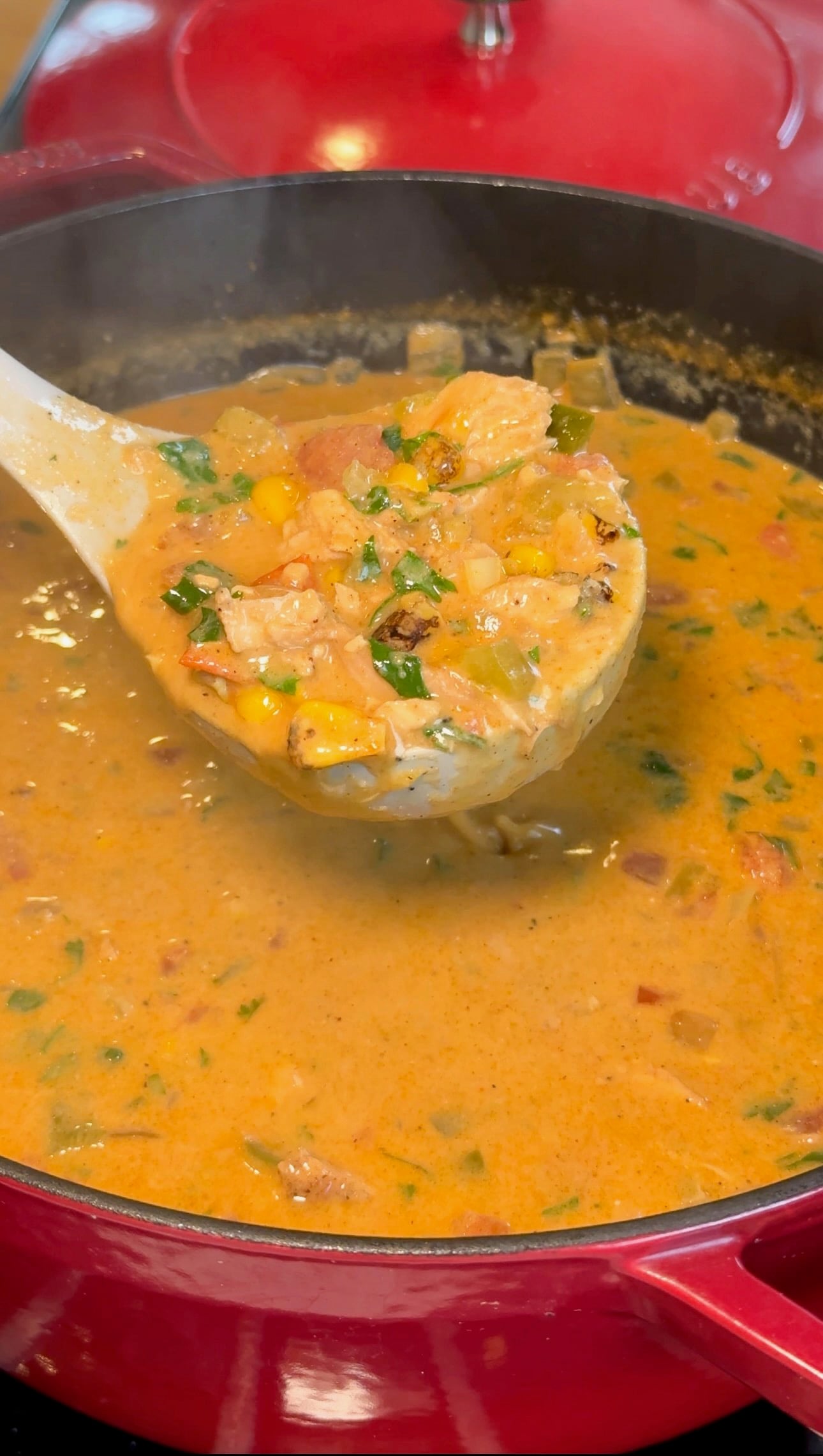 A ladle lifting creamy chicken tortilla soup with chicken, corn, and cilantro out of a red pot.