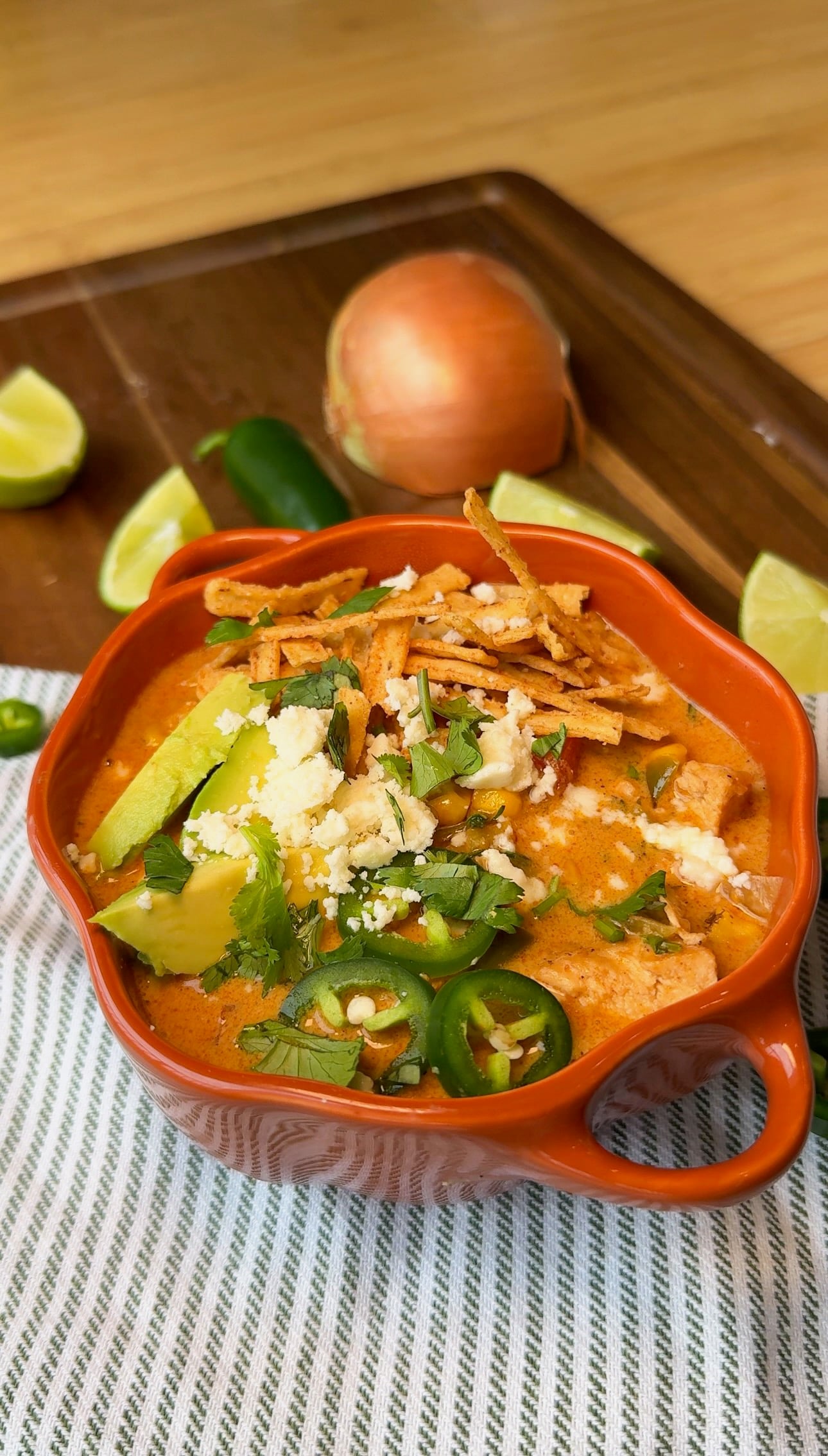 A bowl of creamy chicken tortilla soup topped with avocado, queso fresco, cilantro, and tortilla strips, with limes and a jalapeño.