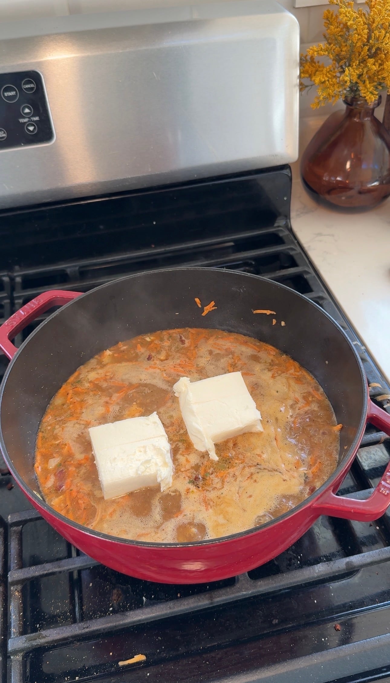 Two blocks of softened cream cheese melting into chicken soup in a red pot.