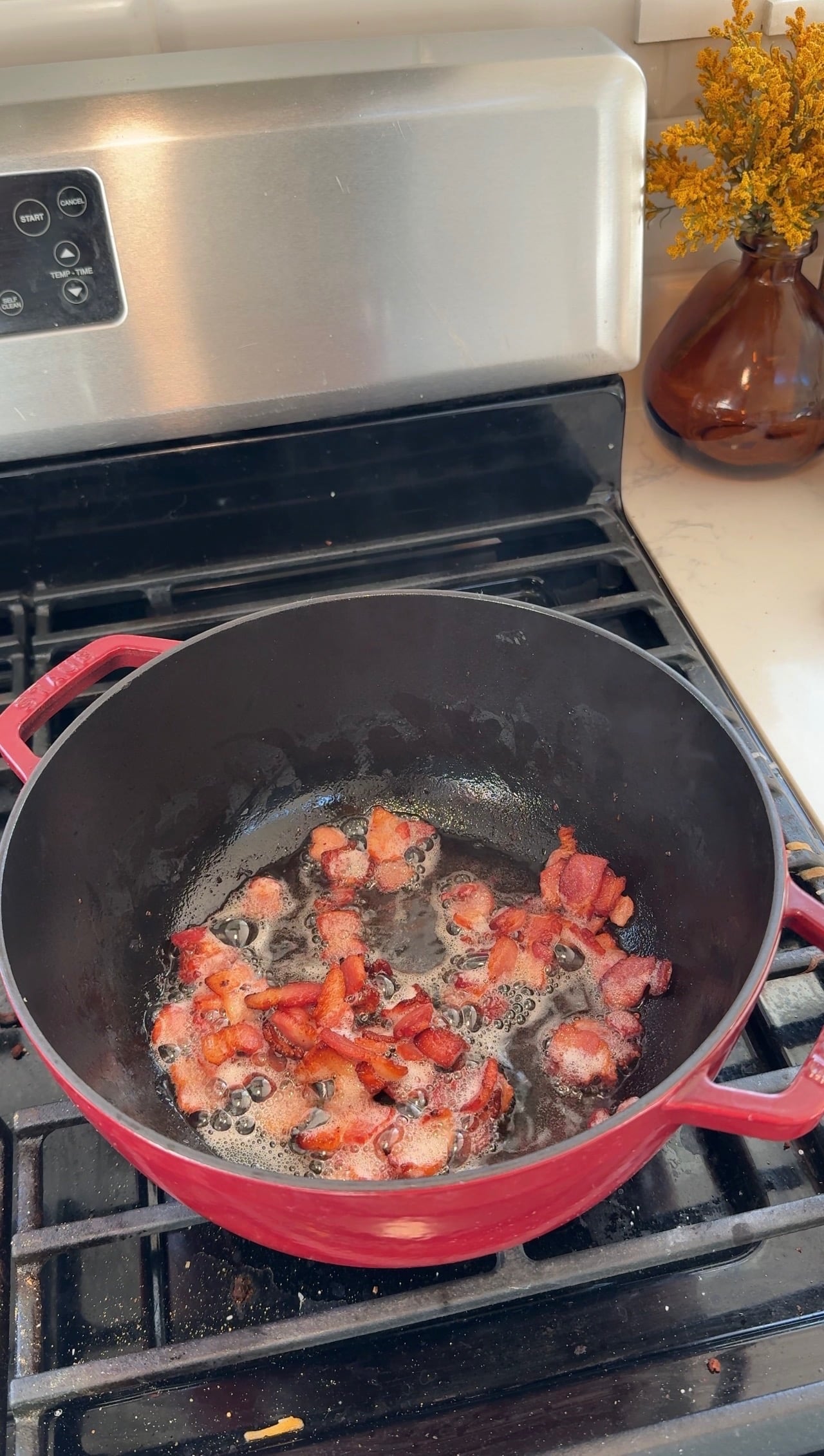 Bacon cooking in the bottom of a red Dutch oven on the stovetop.
