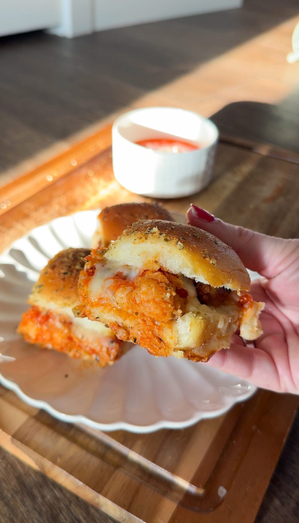 A hand holding a chicken parmigiana slider over a plate with additional sliders and a side of marinara sauce.