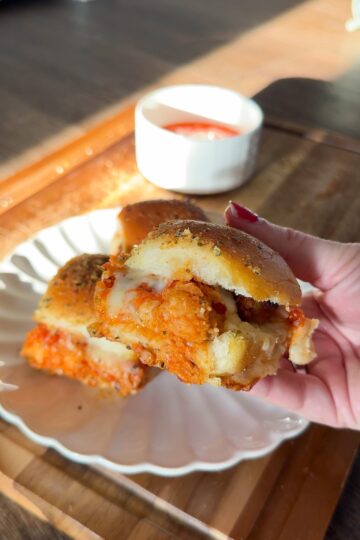 A hand holding a chicken parmigiana slider over a plate with additional sliders and a side of marinara sauce.