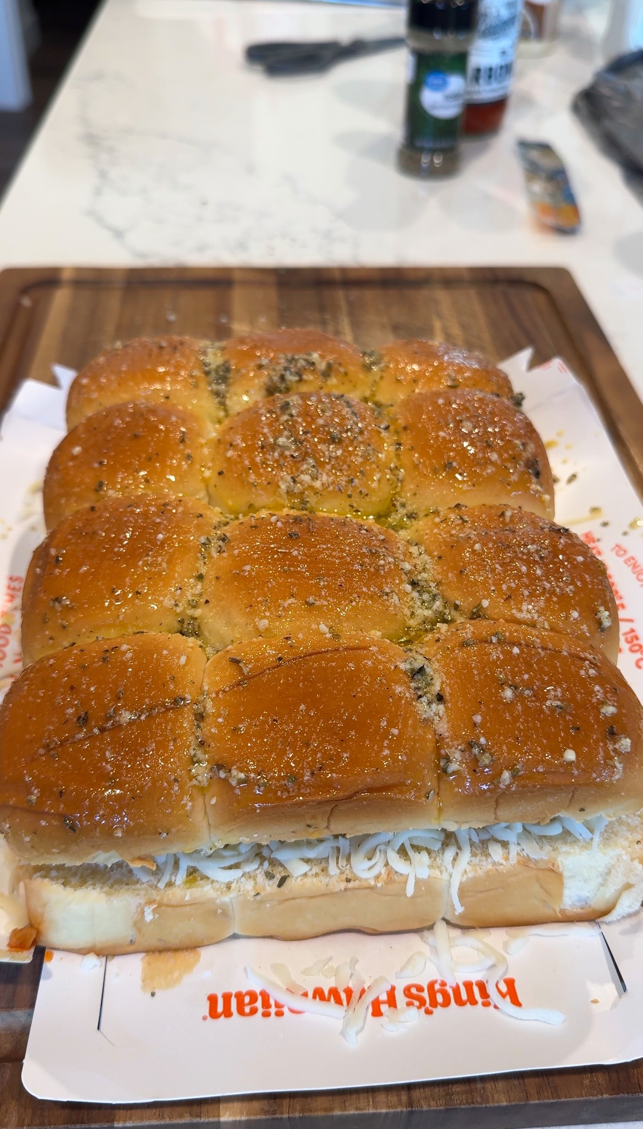 A tray of twelve slider sandwiches on Kings Hawaiian rolls, topped with butter and herbs, sitting on a wooden cutting board.