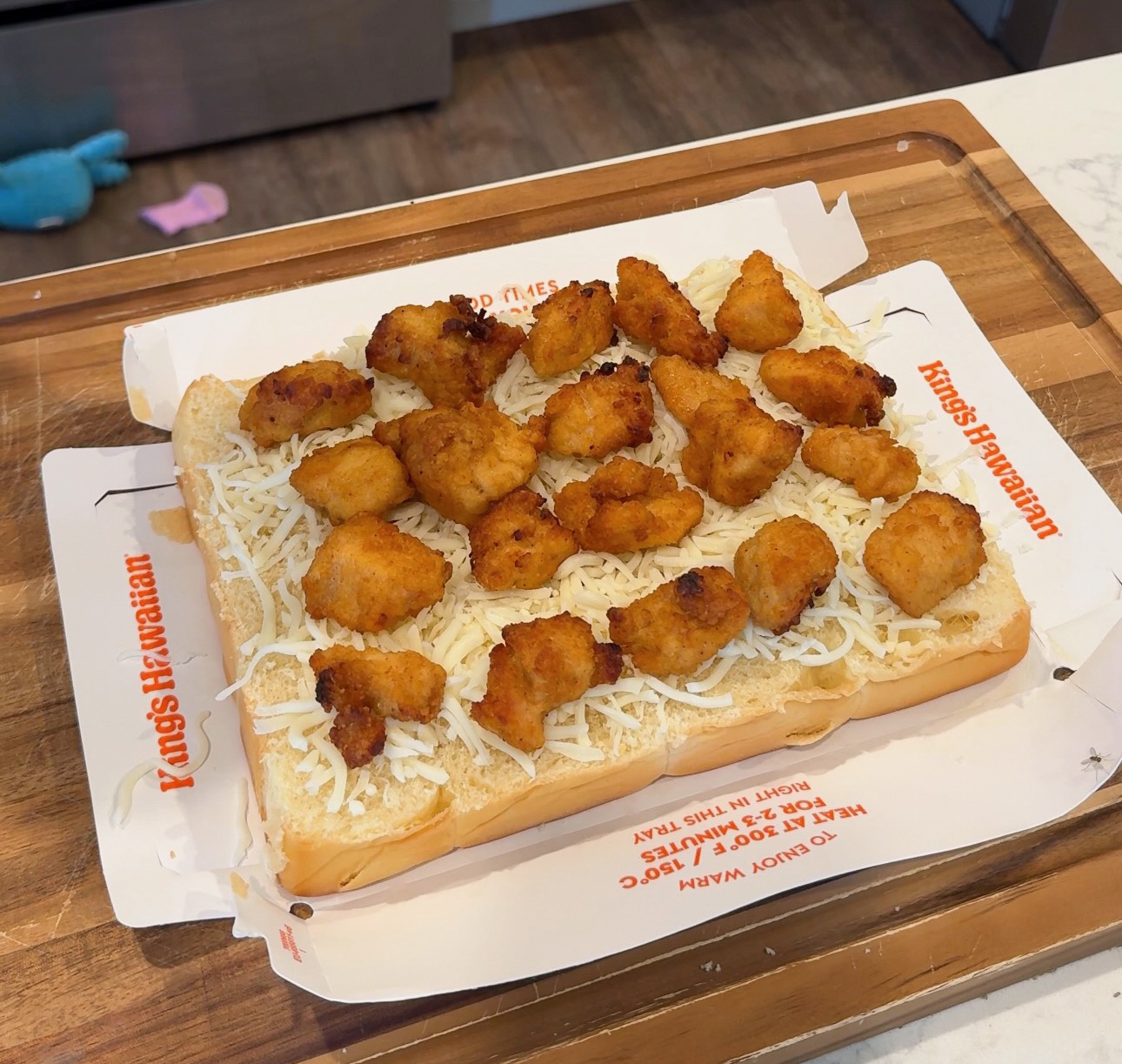 A sheet of Kings Hawaiian sweet rolls topped with shredded mozzarella and bite-sized pieces of fried chicken.