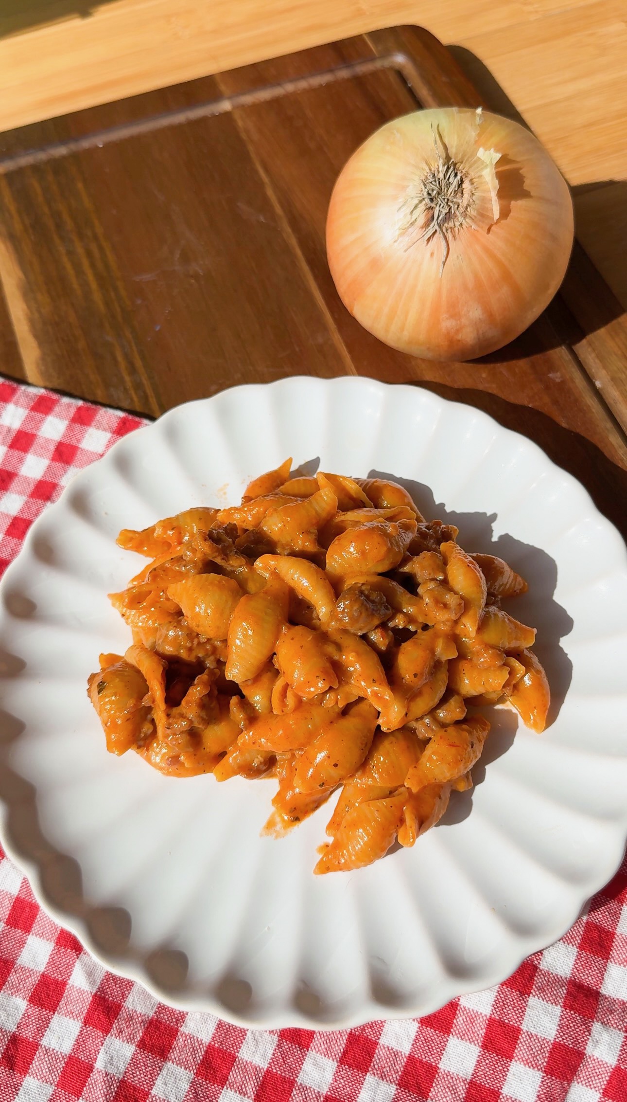 A plate of shell pasta in a creamy red sauce on a red and white checkered cloth next to a whole onion.