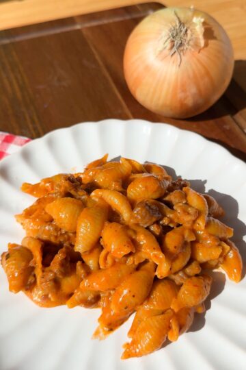 A plate of shell pasta in a creamy tomato and meat sauce, with a whole onion nearby.