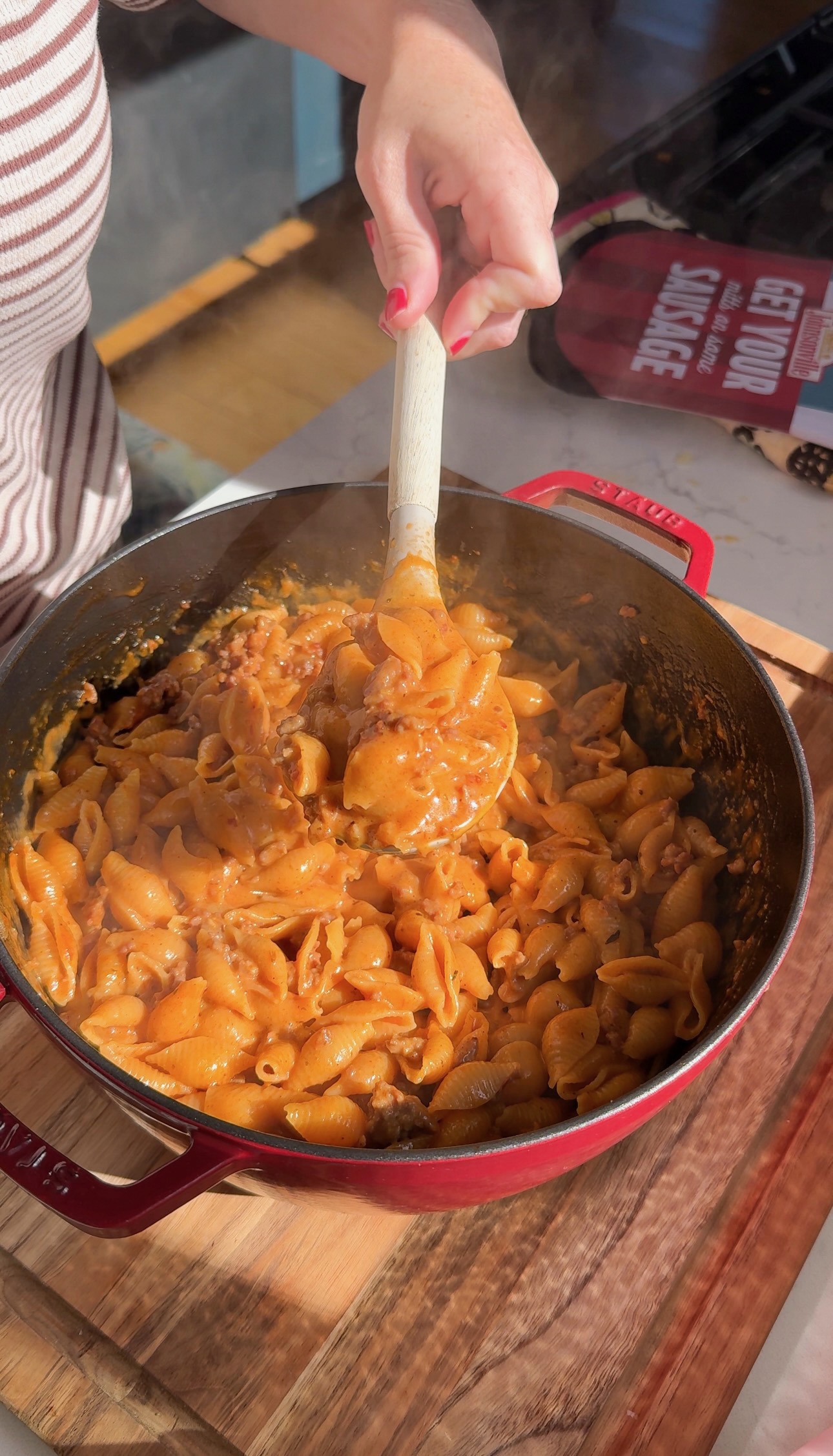 A person lifts a spoonful of creamy pasta shells with meat sauce from a red enameled cast iron pot.