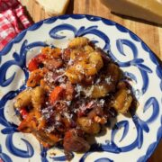 A blue and white patterned plate of gnocchi with sliced sausage and peppers, topped with grated cheese, next to two wedges of parmesan on a wooden board.