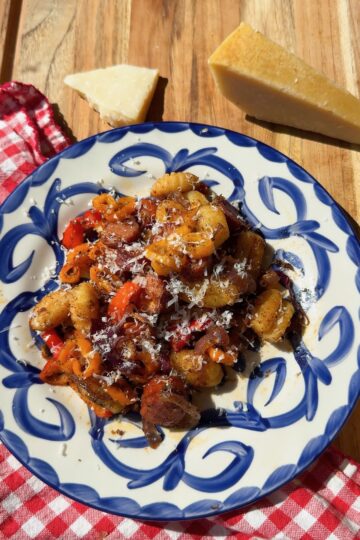 A blue and white patterned plate of gnocchi with sliced sausage and peppers, topped with grated cheese, next to two wedges of parmesan on a wooden board.