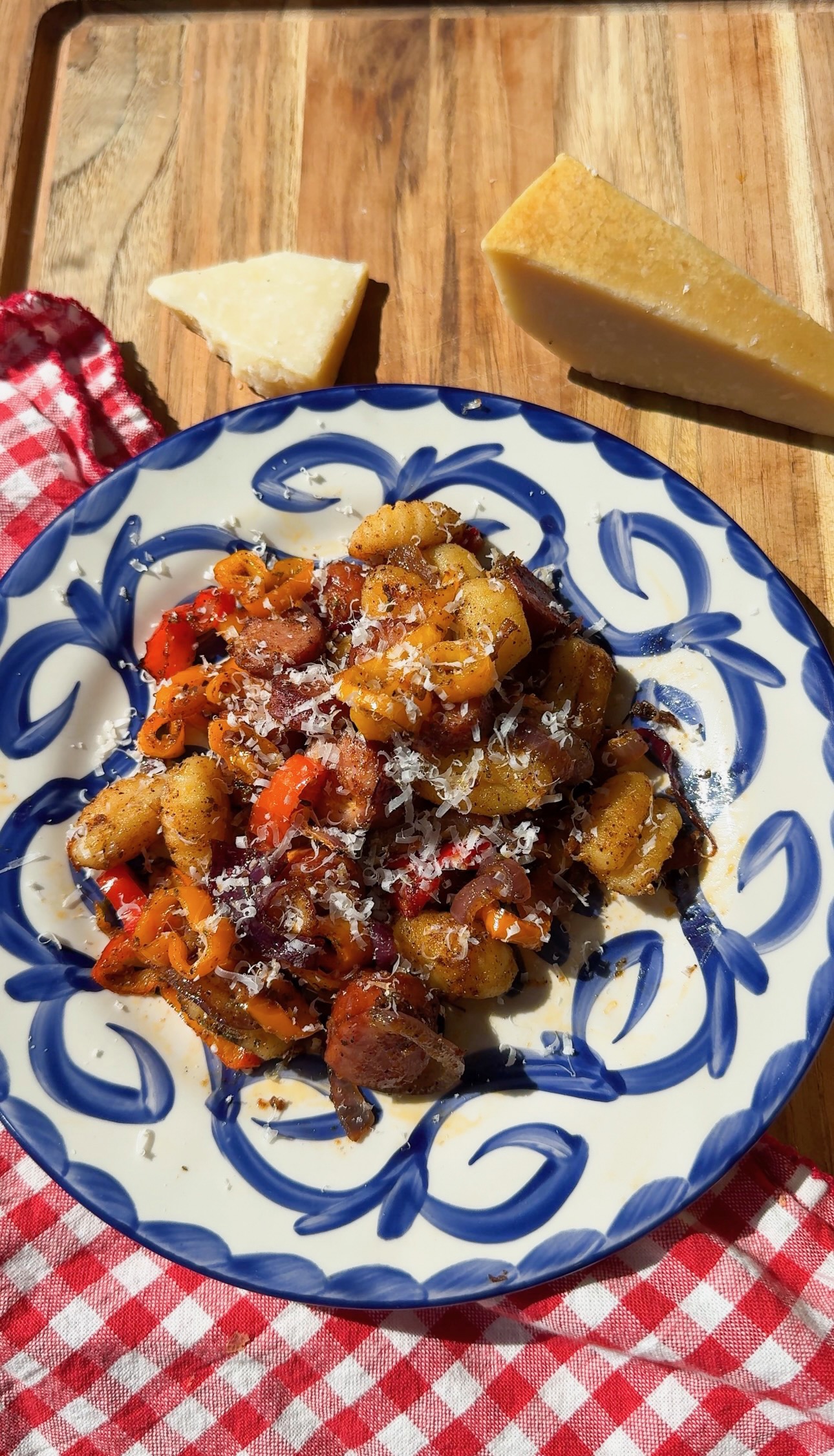 A blue and white patterned plate of gnocchi with sliced sausage and peppers, topped with grated cheese, next to two wedges of parmesan on a wooden board.