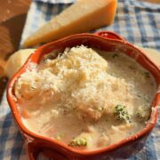 A bowl of creamy pasta with broccoli, topped with grated cheese, with a wedge of cheese in the background.