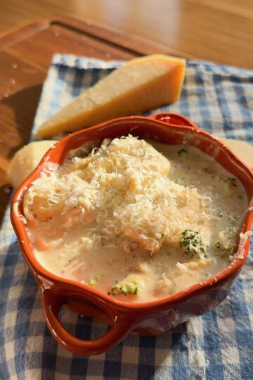 A bowl of creamy pasta with broccoli, topped with grated cheese, with a wedge of cheese in the background.