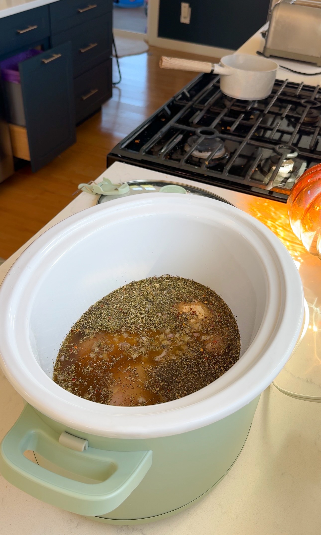 A slow cooker containing chicken breast submerged in broth and topped with a thick layer of dried herbs.