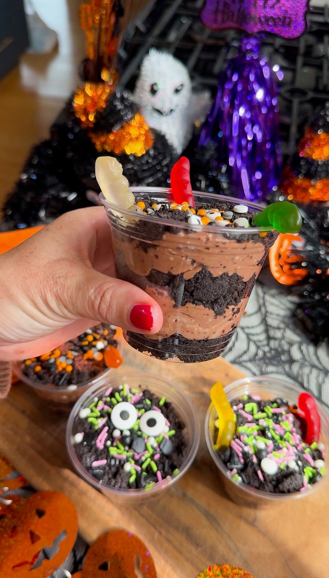A hand holding a chocolate pudding cup topped with gummy worms and Oreo crumbs, with Halloween decorations behind it.