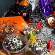 Four Halloween dirt cups layered with chocolate pudding and crushed Oreo dirt, topped with gummy worms and Halloween sprinkles on a wooden board.