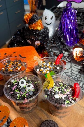 Four Halloween dirt cups layered with chocolate pudding and crushed Oreo dirt, topped with gummy worms and Halloween sprinkles on a wooden board.
