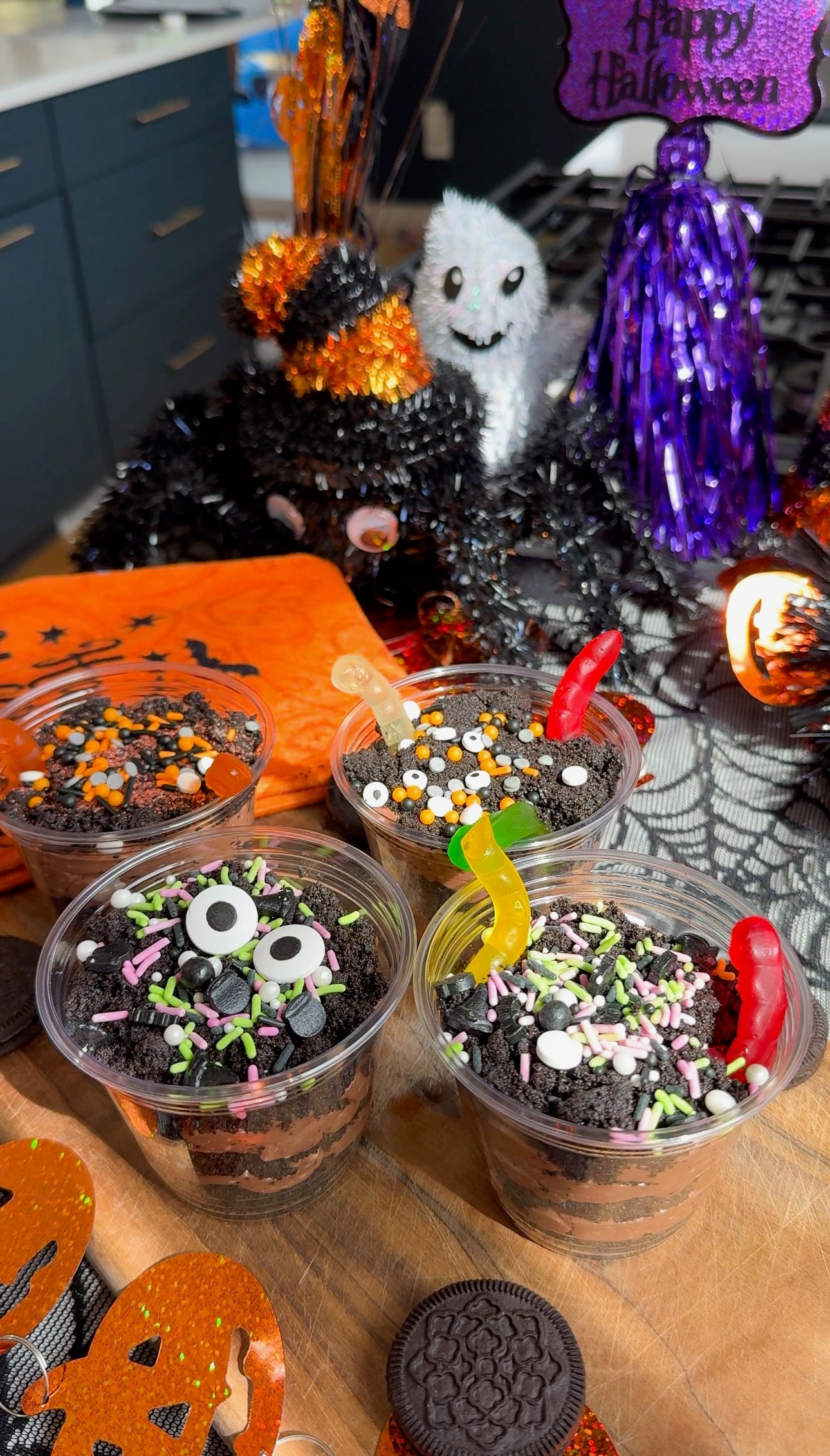 Four Halloween dirt cups layered with chocolate pudding and crushed Oreo dirt, topped with gummy worms and Halloween sprinkles on a wooden board.